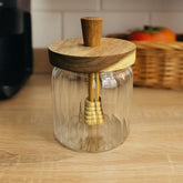 Recipiente de vidrio con líneas verticales, tapa de madera natural y paleta dosificadora para miel.